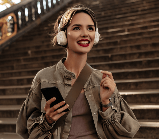 happy-woman-with-short-hair-red-lips-headphones-smiles-woman-jacket-light-pants-holds-phone-outdoors